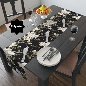 May include: Black and white table runner with a floral and bird design. The runner is made of polyester and features white magnolia flowers, gold accents, and white birds on a black background.