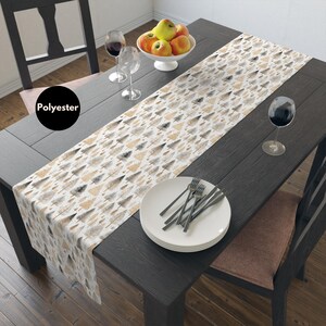 Puede incluir: Un camino de mesa de poliéster con un patrón repetido de árboles estilizados en tonos beige, marrón y gris. El camino de mesa está colocado sobre una mesa de madera oscura, con platos, cubiertos y copas de vino. Un cuenco de manzanas está en el centro.
