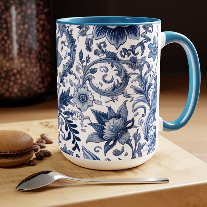 May include: A white ceramic mug with a blue floral pattern and a blue handle. The mug features a detailed design of flowers and leaves in shades of blue. The interior of the mug is also blue. A spoon and a pastry are on a wooden surface.