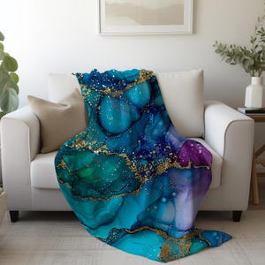 May include: A soft blue, green, and purple throw blanket with gold glitter accents. The blanket has an abstract, marbled design.