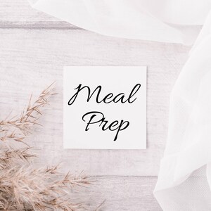 May include: A white card with the text "Meal Prep" written in black cursive script. The card is on a light wood surface with a dried grass plant in the foreground.