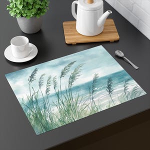 Op de afbeelding: Een blauwe en groene aquarel van hoog gras dat in de wind waait op een strand met een blauwe zee op de achtergrond.
