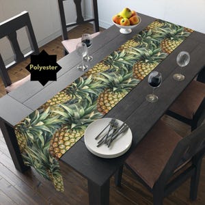 May include: A green and yellow pineapple patterned table runner on a dark wood table with white plates and wine glasses. The table runner is made of polyester.