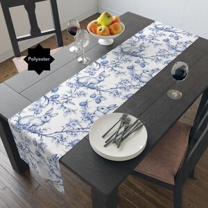 May include: A white table runner with a blue and white pattern of bunnies and flowers. The runner is made of polyester and is 14 inches wide. The runner is on a wooden table with a bowl of apples, two wine glasses, and a set of silverware.