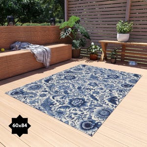 Puede incluir: Una alfombra de exterior con estampado floral azul y blanco con una etiqueta de tamaño de 60x84 pulgadas. La alfombra está colocada en una terraza de madera con un banco y plantas en el fondo.