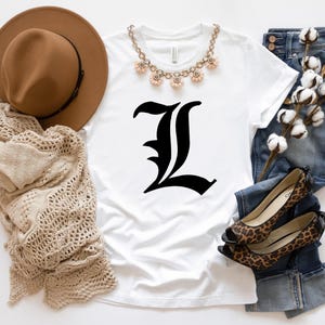 May include: A white t-shirt with a large black letter "L" in a gothic font. The shirt is styled with a brown hat, a beige scarf, a floral necklace, blue jeans, and leopard print shoes.