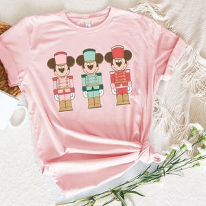 May include: A light pink t-shirt featuring three cartoon nutcracker figures with Mickey Mouse ears. Each figure is dressed in a different color uniform: pink, teal, and red. The shirt is knotted at the bottom and has rolled-up sleeves.