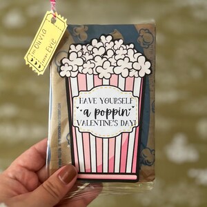 May include: A pink and white striped popcorn bucket with the text "Have yourself a poppin' Valentine's Day!"  A yellow ticket with the text "To Olivia from Evie" is attached to the top of the bucket.