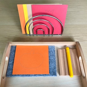 Könnte beinhalten: Ein Holztablett mit einer grauen Filzmatte, orangefarbenem Papier mit einer Kürbiskontur und einem gelben Stift. Ein Metallgestell hält farbiges Papier mit Fruchkonturen. Die Artikel sind auf einer hellen Holzoberfläche angeordnet.
