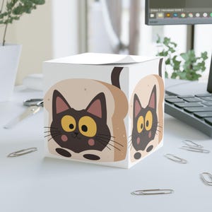 May include: A white cube-shaped notepad dispenser with a cartoon cat design. The cat is brown with yellow eyes and is set against a background of toasted bread. The design is repeated on multiple sides of the dispenser.