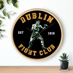 Könnte beinhalten: Runde Wanduhr mit Holzrahmen. Das schwarze Zifferblatt zeigt die Worte "DUBLIN FIGHT CLUB" in Gold. Ein Boxer in grünen Shorts und Handschuhen befindet sich in der Mitte. Das Jahr "1916" ist weiß gedruckt.