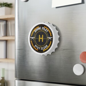 May include: A white bottle cap magnet with a black and yellow design. The design features the words "DRONE PILOT" and "STAY CLEAR" around a central "H" and drone silhouettes. The magnet is attached to a stainless steel surface.