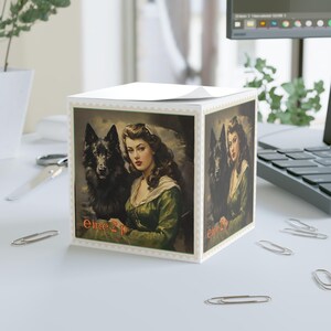 May include: A white cube-shaped notepad featuring a vintage-style portrait of a woman with a black dog. The woman wears a green dress. The notepad has a scalloped border and the text "Eine Z" on the front.