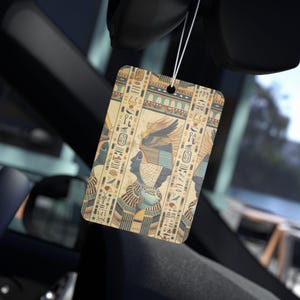 May include: Rectangular car air freshener with an Egyptian hieroglyphic design. The air freshener has a beige background with blue, gold, and black illustrations of pharaohs and hieroglyphs. It is suspended by a white string.