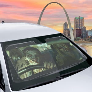 May include: A car windshield sunshade with a graphic of two zombies driving. The sunshade is black and has a gray border. The zombies are wearing casual clothing and have a menacing expression.
