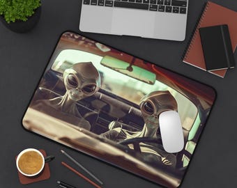 May include: A black mouse pad with a graphic of two gray aliens driving a car. The aliens have large eyes and are smiling. The car is driving down a road with a blurry background.