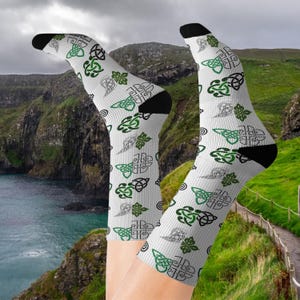 Puede incluir: Calcetines blancos con talones y puntas negras, con un patrón repetido de diseños de nudos celtas verdes y negros. Los calcetines se muestran sobre un fondo de colinas verdes y agua.