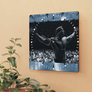 May include: Square wall clock featuring a black and white image of a bodybuilder with arms raised. The clock has a blue border and white markings for the hours. The clock hands are black.