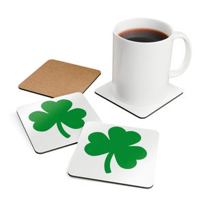 May include: A white ceramic mug filled with dark liquid sits on a square white coaster with a black trim. Three square coasters with a white background and a green shamrock design are also present, along with a cork coaster.
