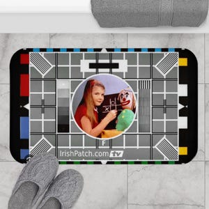 May include: Rectangular bath mat featuring a retro TV test pattern design with a central image of a girl and a clown puppet. The mat has a black border and the text "IrishPatch.com/tv". Gray slippers are in the foreground.