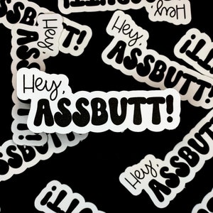 Könnte beinhalten: Eine Sammlung weißer Aufkleber mit schwarzer Beschriftung. Die Aufkleber sagen "Hey, ASSBUTT!" in verschiedenen Schriftarten und Größen.