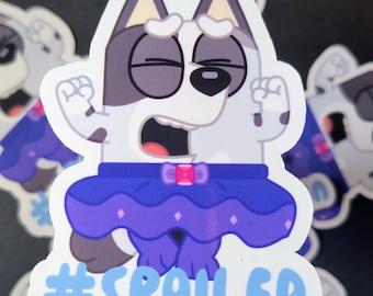 May include: A sticker featuring a cartoon dog with gray and white fur, wearing a blue skirt with a pink gem. The dog has its eyes closed and mouth open, with the hashtag #SPOILED in blue text.