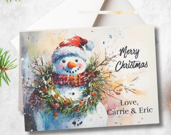 May include: A watercolour Christmas card featuring a snowman wearing a red and white hat and a red and white scarf. The snowman is surrounded by a wreath of green branches and red berries. The card reads "Merry Christmas" and "Love, Carrie & Eric".