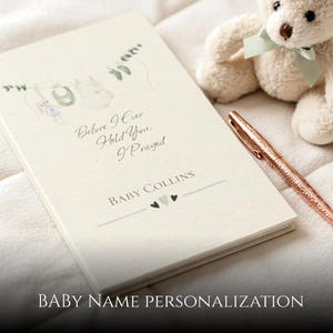 Puede incluir: Un libro de recuerdos para bebés de color crema con el texto "Before I Ever Held You, I Prayed" y "Baby Collins". Un bolígrafo de oro rosa y un osito de peluche con un lazo verde están al lado del libro. El libro tiene una ilustración de acuarela de ropa de bebé.