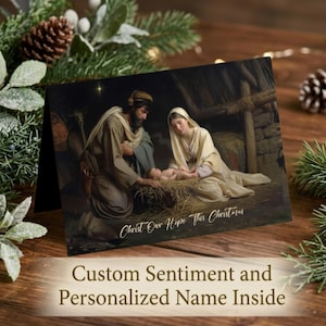 May include: A folded greeting card featuring a nativity scene with a man and woman kneeling beside a baby in a manger. The text Christ Our Hope This Christmas is printed in white script at the bottom. The card is surrounded by pine branches.