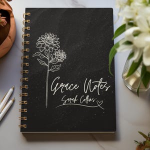 May include: Black spiral bound notebook with a white floral design and the text "Grace Notes Sarah Collins" on the front cover.