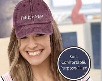 Puede incluir: Gorra de béisbol granate con el texto "Faith > Fear" bordado en blanco. La gorra está hecha de un material suave y tiene una visera curva. La imagen también incluye un gráfico circular con las palabras "Soft. Comfortable. Purpose-Filled."
