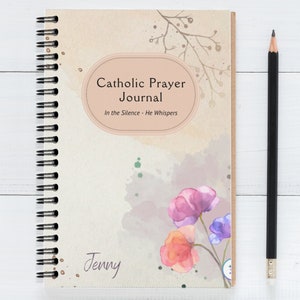 May include: A beige spiral bound journal with a watercolor design of flowers. The cover has the text "Catholic Prayer Journal" and "In the Silence - He Whispers". The name "Jenny" is written on the bottom of the cover.