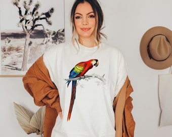 May include: White t-shirt featuring a colorful illustration of a parrot perched on a branch. The parrot has red, yellow, blue, and green feathers. The shirt is paired with a brown jacket and a tan hat.
