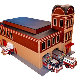 May include: A miniature fire station model featuring a brown and beige brick exterior with a tall tower, a flat roof, and multiple arched bays. Three small fire engine trucks are parked inside and outside the structure on a gray base.
