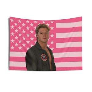 May include: A pink American flag tapestry with a portrait of a man in a green flight suit. The flag design features white stars on a pink field and pink and white stripes.