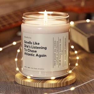 Puede incluir: Una vela de vidrio transparente en un frasco con una etiqueta beige que dice "StarGirls Smells Like She's Listening To Chase Atlantic Again". La vela está encendida y la llama es visible. La etiqueta también incluye el texto "Vela de soja hecha a mano. Elaborada en California 50-60 horas de tiempo de combustión 9 oz Mantener alejado de las llamas."