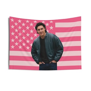 May include: A pink American flag tapestry with a person in a dark jacket and jeans. The flag design features pink stripes and white stars. The person is smiling and has their hands in their pockets.