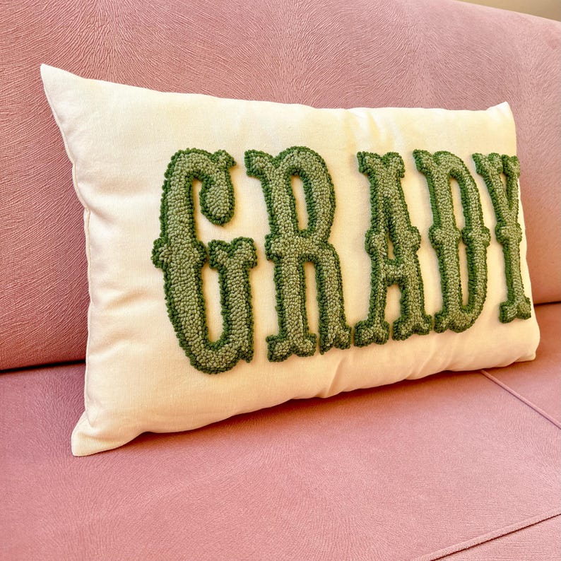 Puede incluir: Coj&iacute;n rectangular crema con el nombre "GRADY" bordado en verde, con letras de bloque. El coj&iacute;n est&aacute; colocado sobre un sof&aacute; rosa. Las letras texturizadas destacan sobre el fondo liso del coj&iacute;n.