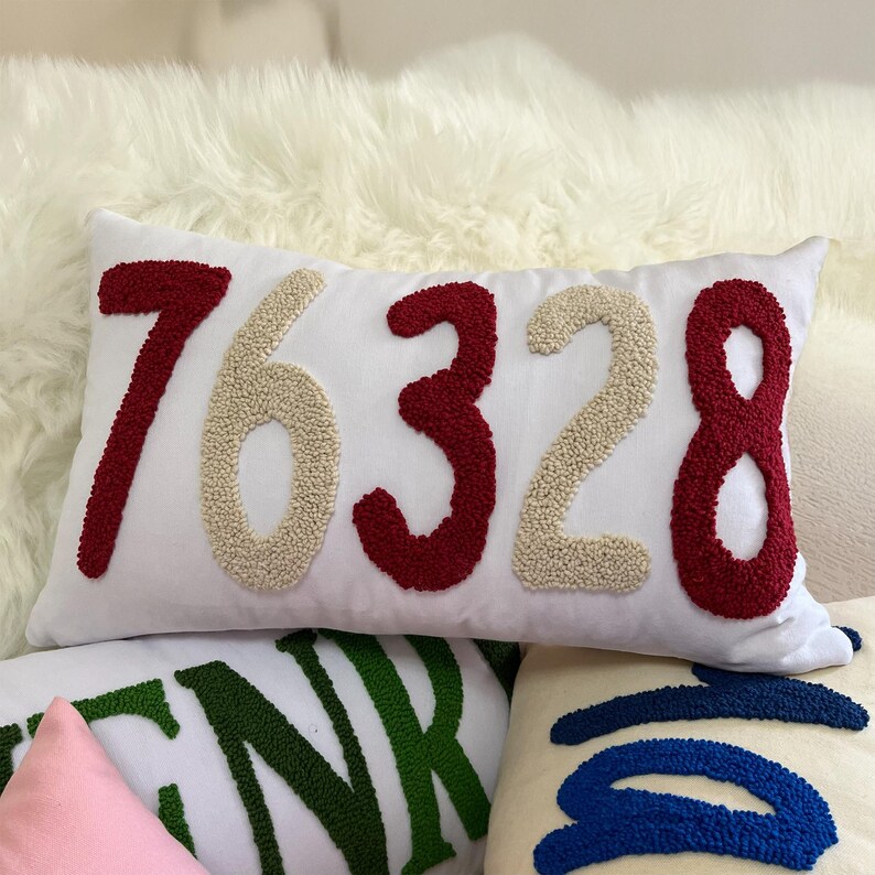 May include: White rectangular cushion embroidered with the number "76328" in red and beige. The cushion is on a white fluffy surface, with other cushions visible in the background.