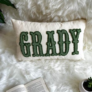 Puede incluir: Coj&iacute;n rectangular color crema con el nombre "GRADY" bordado en letras verdes estilizadas. El coj&iacute;n est&aacute; colocado sobre una superficie de piel sint&eacute;tica blanca y esponjosa. Un libro y una peque&ntilde;a planta en maceta son visibles en el fondo.
