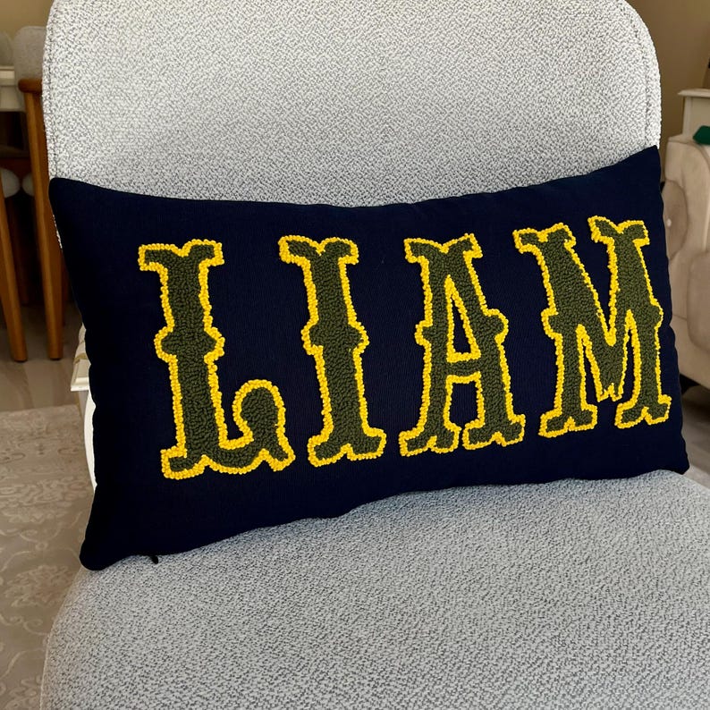 Boho home decor style punch needle pillow featuring the name Liam in yellow embroidery on a navy base, displayed on a modern textured chair.