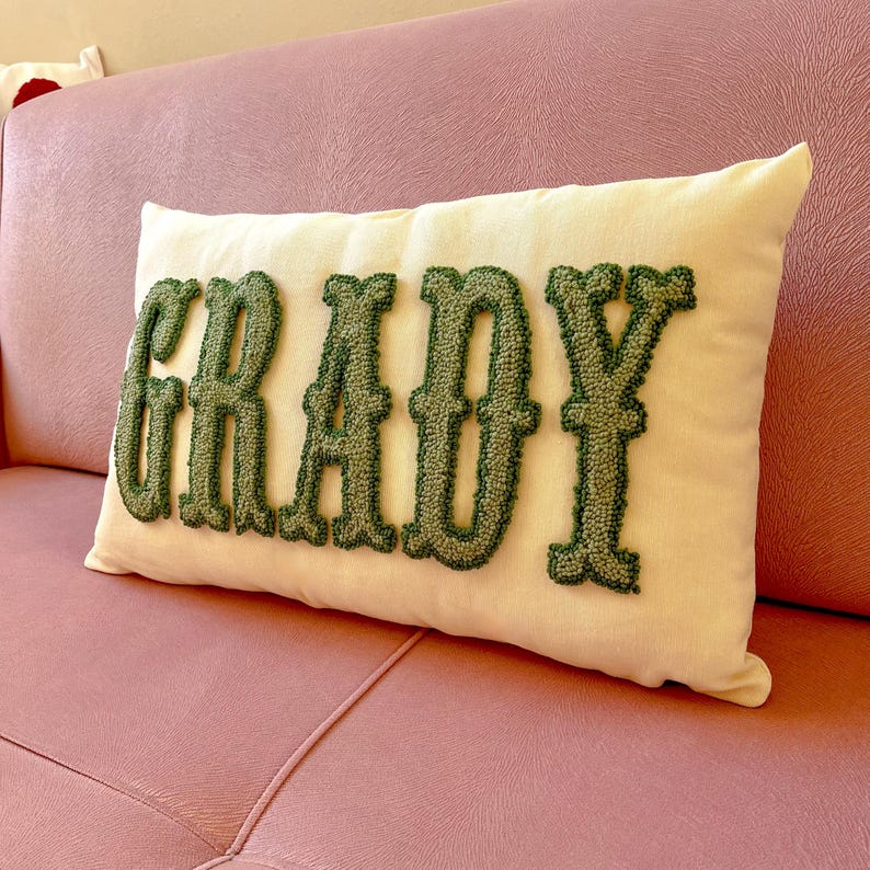 Puede incluir: Un coj&iacute;n rectangular crema con el nombre "GRADY" bordado en letras verdes estilizadas. El coj&iacute;n est&aacute; colocado sobre un sof&aacute; rosa. Las letras son en relieve, dando una apariencia texturizada.