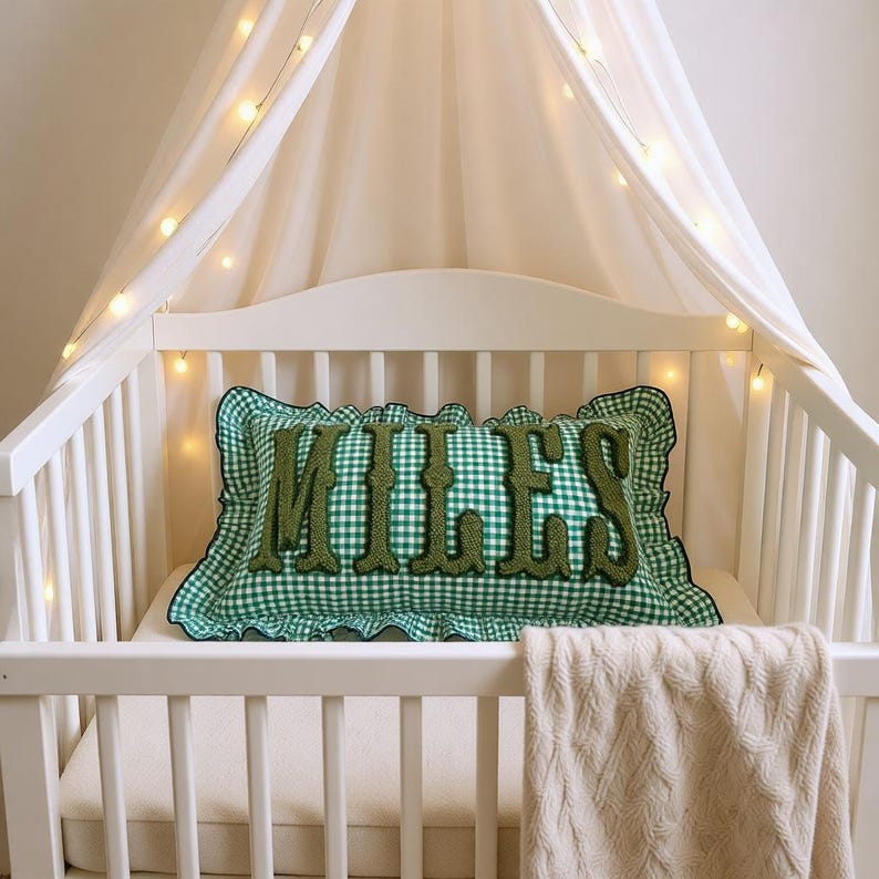 May include: A white cot with a green and white gingham pillow embroidered with the name "MILES" in green letters. The pillow has a ruffled edge. A white canopy with fairy lights is above the cot, and a cream-coloured blanket hangs over the side.
