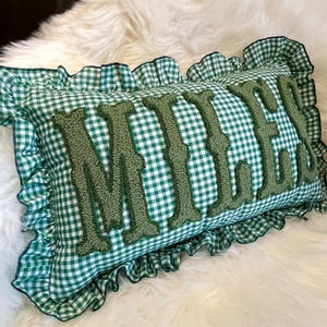 May include: A rectangular cushion with a green and white gingham pattern. The word "MILES" is embroidered in a textured green font across the centre. The cushion has a ruffled edge in the same gingham fabric.