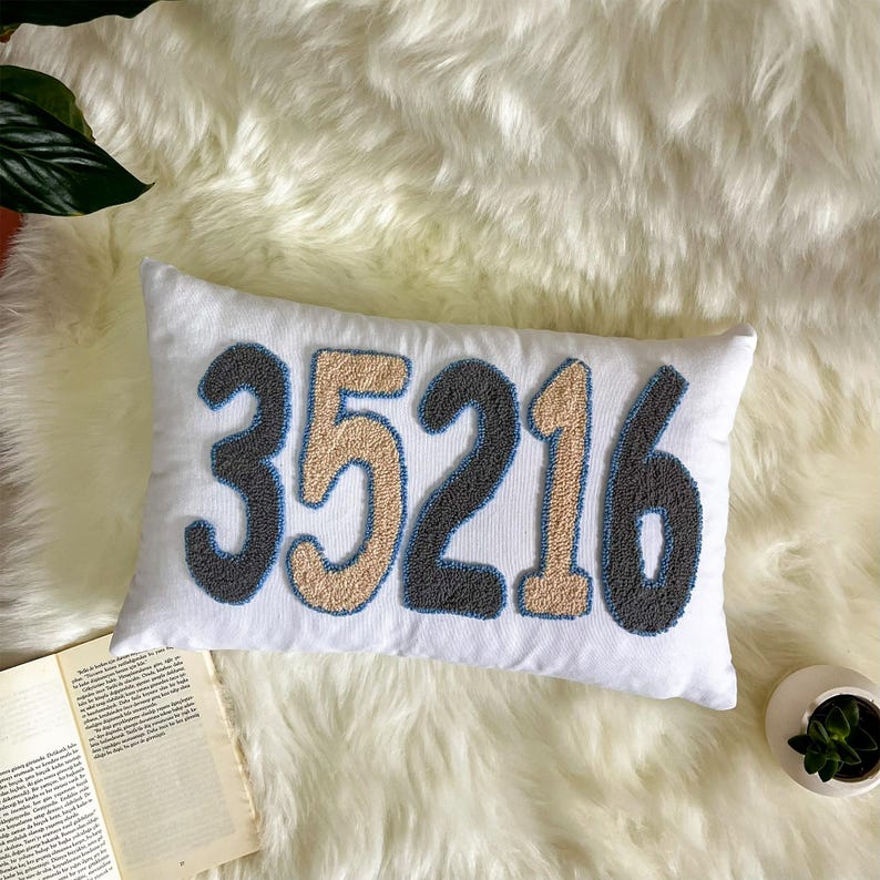 May include: A white rectangular cushion embroidered with the numbers "35216" in grey, beige, and blue. The cushion is displayed on a fluffy white surface, suitable for home decor.