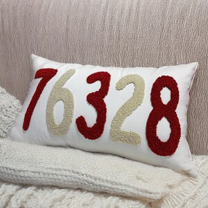 May include: A white rectangular cushion embroidered with the numbers 76328 in red and beige. The cushion rests on a white knitted blanket. The background is a textured, light-coloured surface.