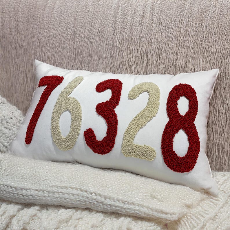 May include: A white rectangular cushion embroidered with the numbers 76328 in red and beige. The cushion rests on a white knitted blanket. The background is a textured, light-coloured surface.