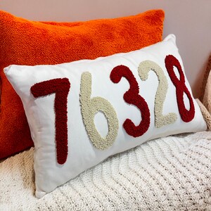 May include: White rectangular cushion with the number "76328" in red and beige textured fabric. An orange cushion is behind it. The cushions are on a white knitted blanket. Home decor.