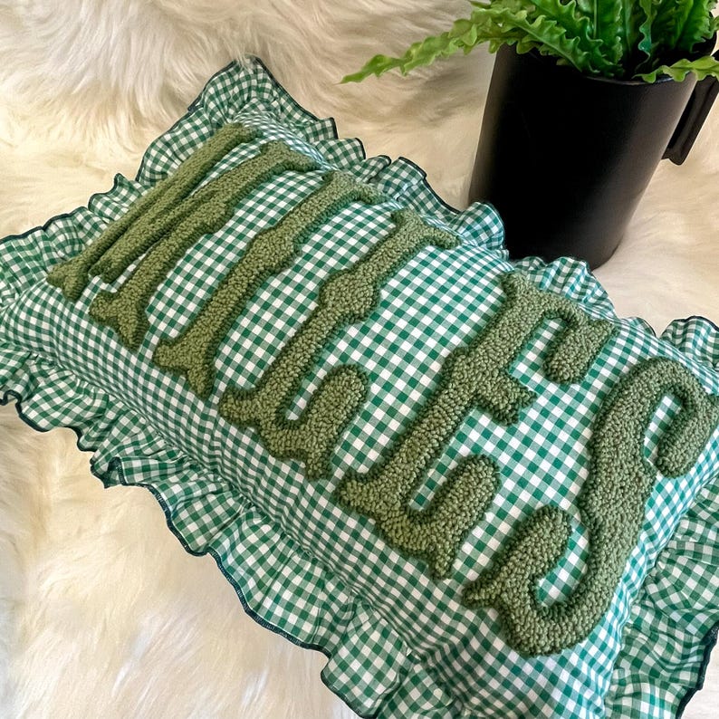 May include: A rectangular cushion with a green and white gingham pattern and a frilled edge. The word "little" is embroidered in a textured green thread across the centre of the cushion. A black pot with a green plant is in the background.