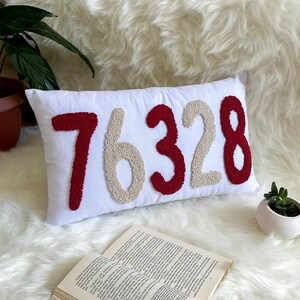 May include: White rectangular cushion featuring the number "76328" embroidered in red and beige. The cushion is on a white fluffy surface, with a plant and a book in the background.